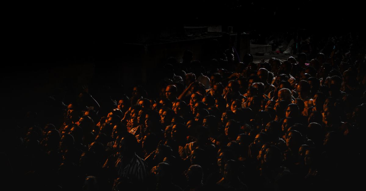A crowd of dark silhouettes with one figure lit by a single gold light